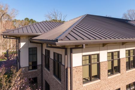 An elevated, high-resolution drone shot showcasing the sleek, standing seam metal roof of a modern commercial building. The image highlights the clean lines, sharp angles, and durable, dark gray finish of the metal roofing system, which provides superior weather resistance and a contemporary architectural look.