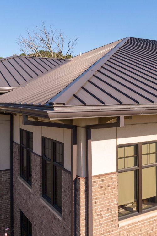 An elevated, high-resolution drone shot showcasing the sleek, standing seam metal roof of a modern commercial building. The image highlights the clean lines, sharp angles, and durable, dark gray finish of the metal roofing system, which provides superior weather resistance and a contemporary architectural look.